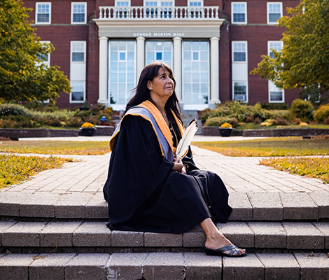 A Mi'kmaq Maliseet Bachelor of Social Work graduate sitting in the courtyard with an eagle feather